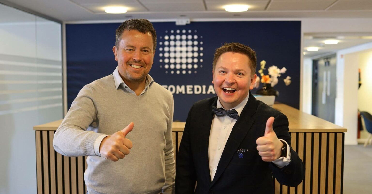 Thomas Vejlemand og Jaakko Haarala smiler og holder tomler opp med Infomedias logo i bakgrunnen
